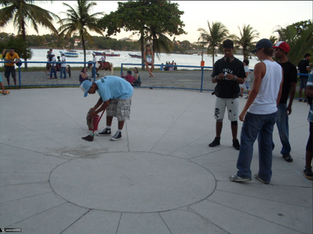 A imagem mostra um grupo de jovens reunidos em uma área aberta, pavimentada e circular, próxima a uma praia ou lagoa. O cenário ao fundo revela o mar, algumas embarcações ancoradas e várias pessoas desfrutando do local, inclusive uma criança montada em um pônei e outras sentadas perto da cerca azul que delimita o espaço.
No primeiro plano, um rapaz de camiseta azul, bermuda xadrez e tênis está agachado, limpando o chão com uma vassoura pequena e um pano. Ele parece concentrado na tarefa. Ao redor dele, quatro outros jovens observam — dois estão de boné, um usa camiseta preta e outro branca, e todos têm um visual casual, típico de um encontro entre amigos.
O ambiente transmite uma atmosfera descontraída, de fim de tarde, com palmeiras, céu claro e pessoas aproveitando o tempo ao ar livre. Tudo indica que se trata de um espaço público à beira-mar, possivelmente usado para lazer, esportes ou convivência.