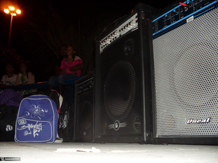 A imagem mostra uma cena noturna ao ar livre, provavelmente durante um evento musical ou ensaio. Em primeiro plano, vemos grandes caixas de som e amplificadores — incluindo um da marca Oneal — dispostos lado a lado sobre o chão de concreto. Perto das caixas há uma mochila azul e branca, com desenhos e a inscrição “Billabong Girls”.
Ao fundo, algumas pessoas estão sentadas em um banco, parcialmente iluminadas pela luz de um poste, que aparece no canto esquerdo superior da foto. Entre elas há uma criança de blusa rosa sentada no colo de um adulto. As árvores e a iluminação sugerem que o local é uma praça ou espaço público, e o clima geral é de preparação ou intervalo de uma apresentação musical.