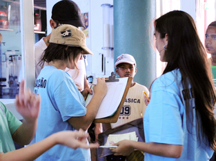 Grupo de crianças e adolescentes interagindo em um ambiente escolar ou comunitário. No centro da imagem, uma menina de boné bege e camiseta azul segura uma prancheta e parece estar preenchendo uma folha com uma caneta. À sua frente, uma garota de cabelo longo também de camiseta azul segura papéis e canetas, parecendo ajudar na organização. Ao fundo, um garoto usando uma camiseta clara e boné branco observa a cena atentamente. Outras crianças aparecem ao redor, algumas olhando para a câmera, outras envolvidas na atividade. O ambiente é claro, com paredes azul claro e uma grande coluna verde. As pessoas parecem estar participando de uma atividade de registro ou cadastramento, talvez para algum evento ou projeto.