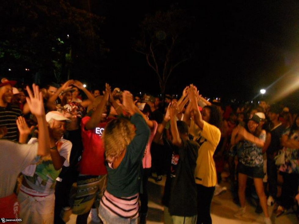 Grupo de pessoas em uma festa ou celebração ao ar livre durante a noite. Muitos parecem estar dançando ou brincando, levantando as mãos e sorrindo. Há uma sensação de alegria coletiva, com roupas casuais e coloridas, e algumas pessoas usam bonés e acessórios descontraídos. Ao fundo, é possível ver mais pessoas participando e algumas árvores, sugerindo que a comemoração acontece em um parque ou praça.

Consultoria de descrição de imagem, feita por Luciana Alvarenga.