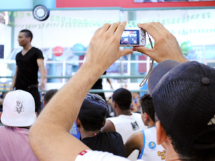 Plateia em um evento esportivo. Um homem usando boné preto está em destaque, segurando uma câmera digital e tirando uma foto ou filmando o que parece ser uma apresentação ou competição. Outras pessoas na plateia também aparecem, algumas usando bonés e camisetas. O foco da câmera está em um homem de camiseta preta no palco, que aparece desfocado à esquerda. O local é claro, com decoração esportiva e bolas coloridas ao fundo, sugerindo que é em uma academia ou ginásio.