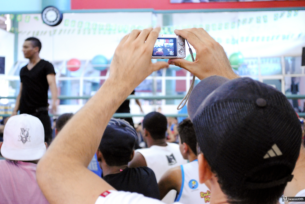 Plateia em um evento esportivo. Um homem usando boné preto está em destaque, segurando uma câmera digital e tirando uma foto ou filmando o que parece ser uma apresentação ou competição. Outras pessoas na plateia também aparecem, algumas usando bonés e camisetas. O foco da câmera está em um homem de camiseta preta no palco, que aparece desfocado à esquerda. O local é claro, com decoração esportiva e bolas coloridas ao fundo, sugerindo que é em uma academia ou ginásio.