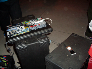 A foto mostra um ambiente externo, provavelmente à noite, onde há dois equipamentos pretos grandes — possivelmente caixas de som ou cases de transporte — usados como apoio para outros objetos.
Sobre uma das caixas, há um notebook fechado coberto por diversos adesivos coloridos (com logotipos, frases e marcas, como “URGH”, “Atmo Imbatível” e outras de estilo urbano, ligadas a skate, música ou cultura alternativa). Do notebook sai um cabo branco, conectado a um iPod clássico (modelo com roda de rolagem branca e tela pequena), que está apoiado na outra caixa.
Há também fios e cabos conectando os dispositivos, sugerindo que o notebook e o iPod estão sendo usados em conjunto, talvez para tocar ou transferir músicas.
Ao redor, é possível ver partes de pessoas — pernas e roupas coloridas — indicando que a cena ocorre em um espaço público, possivelmente em um evento ou encontro musical informal.
A iluminação é artificial, e o chão é de cimento liso.