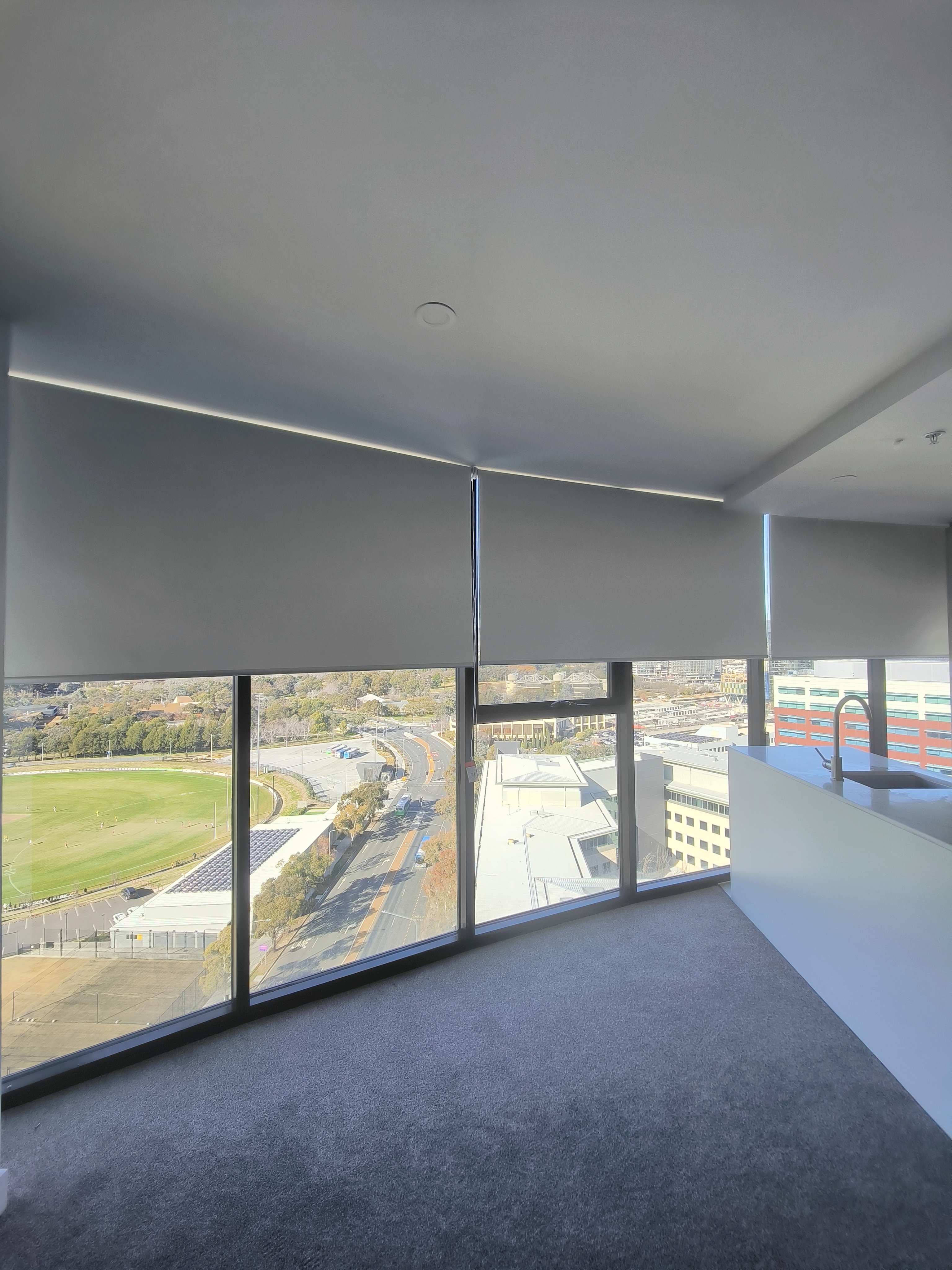 High-rise room with large windows showing a city view. Grey blinds, carpet, and a white counter with a sink create a modern, calm setting.