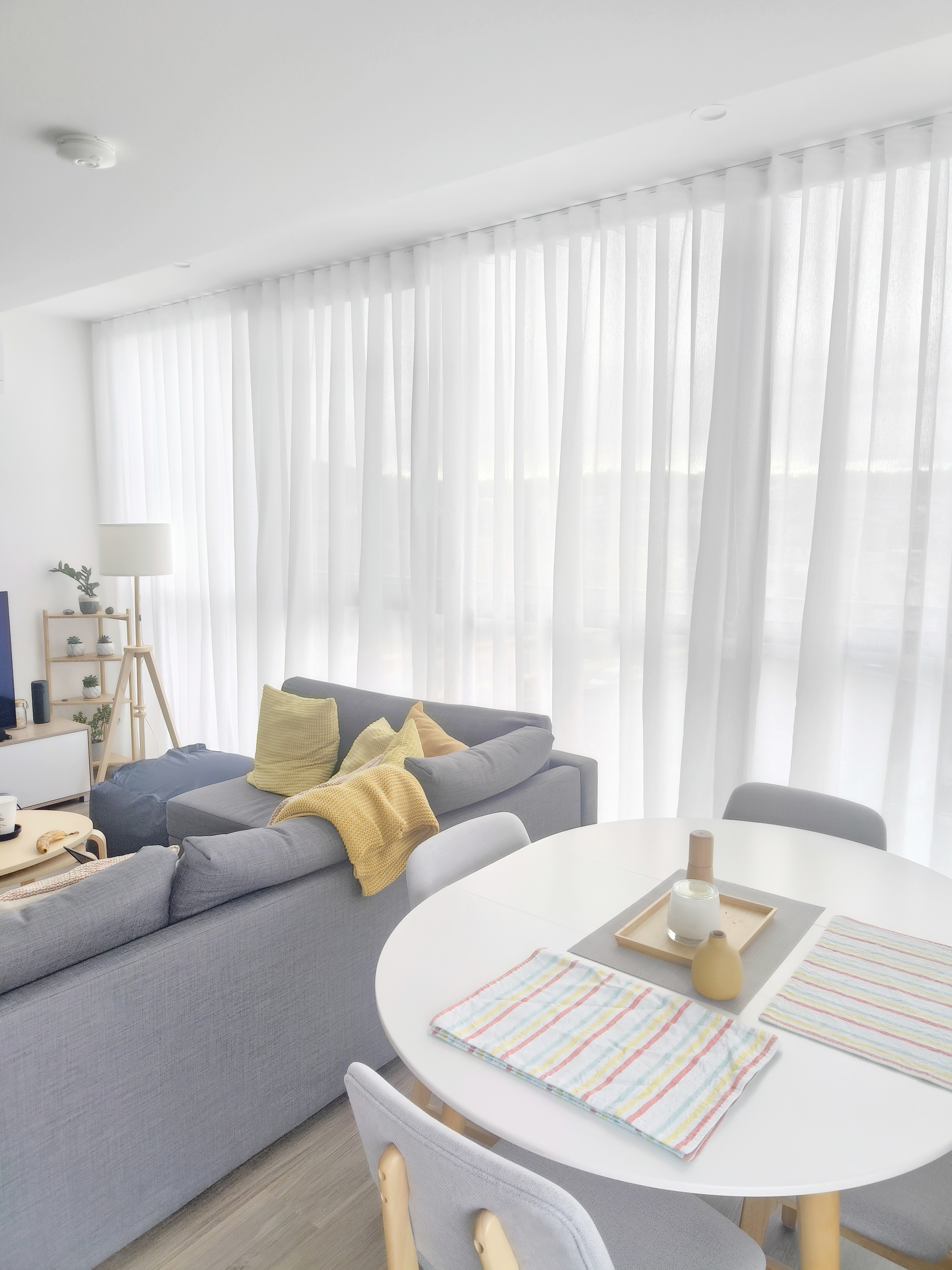 Bright living room with a gray sofa, yellow pillows, and a white table with striped placemats. Soft light from sheer curtains creates a cozy vibe.