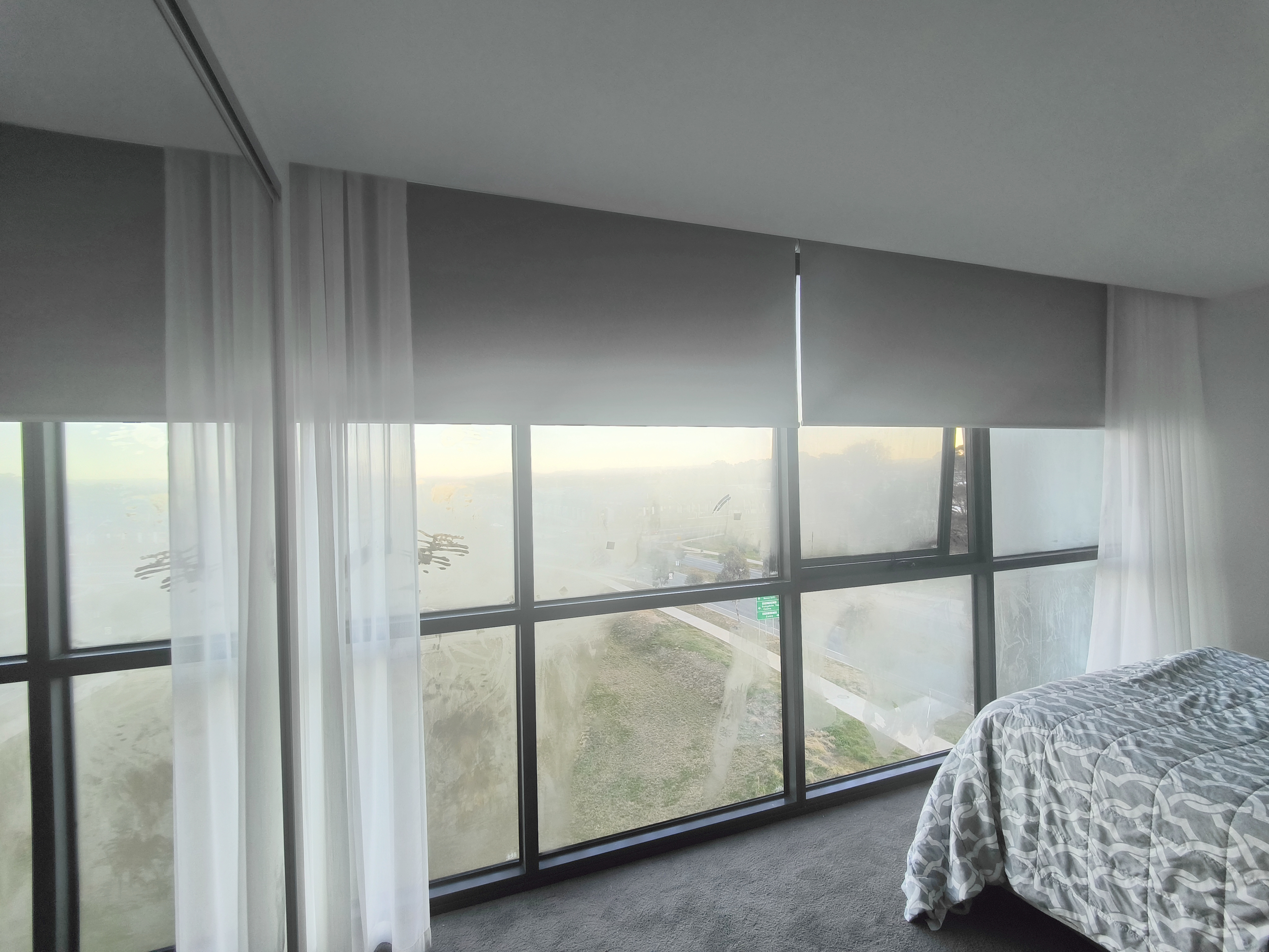 Bedroom with layered sheer curtains and blockout roller blinds over large windows.