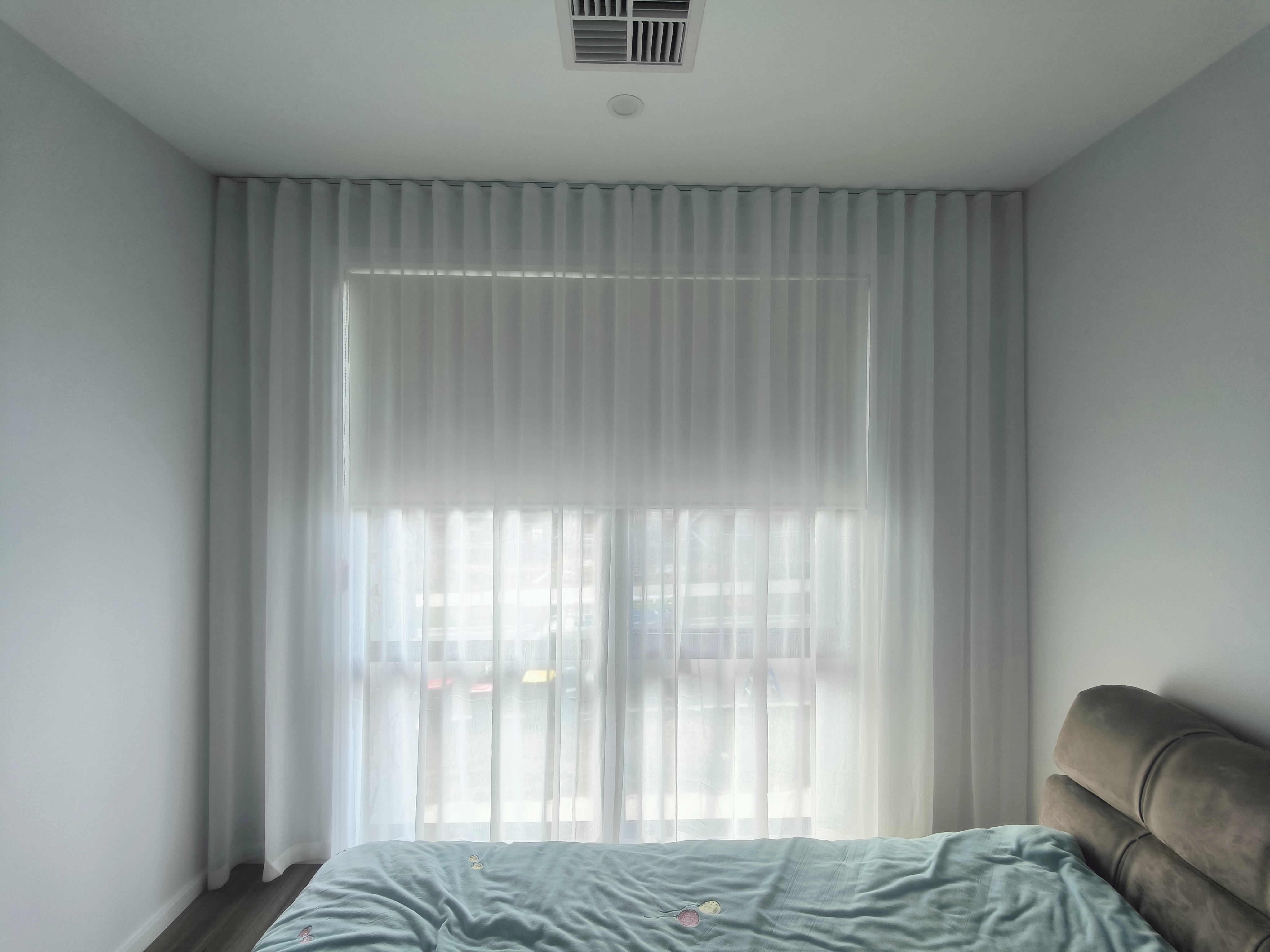 A bedroom with a soft gray bed and light gray comforter. Sheer white curtains cover a large window, creating a calm, bright ambiance.