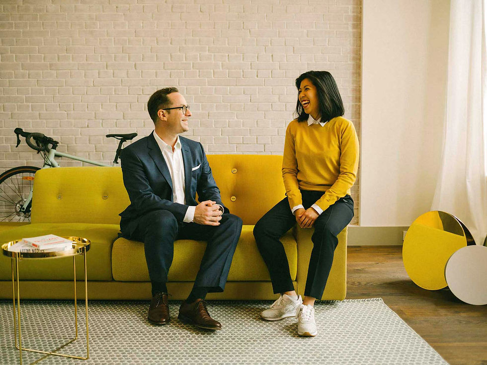 Ein Mann und eine Frau sitzen auf einem gelben Sofa und sehen sich lachend an. Warme gelbe Farben. Teambild einer Personal Branding Serie