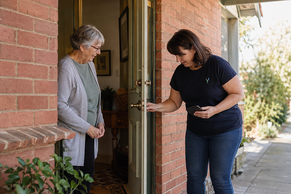 A woman in a dark navy T-shirt at an open front door, greeted by an older woman standing inside.