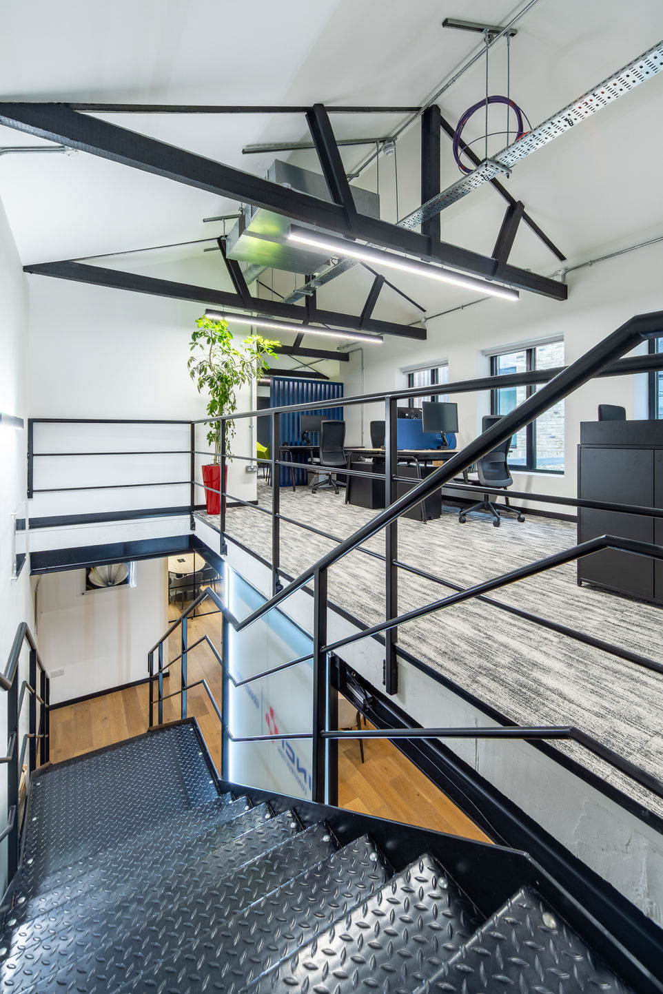 Mezzanine walkway with metal railings overlooking glass offices and workstations below.