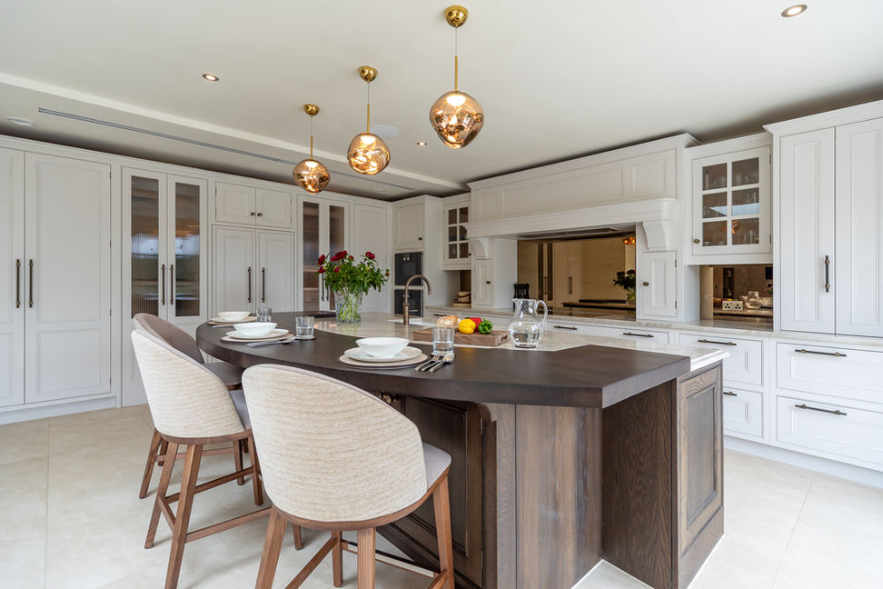 Bright classic kitchen with white cabinetry, dark island worktop and upholstered stools under brass pendant lighting.