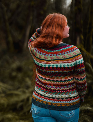 Woman with long red hair wearing a colorful hand knit sweater surrounded by trees and ferns.