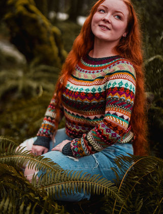 Woman with long red hair wearing a colorful hand knit sweater surrounded by trees and ferns.
