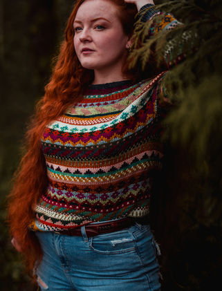 Woman with long red hair wearing a colorful hand knit sweater surrounded by trees and ferns.