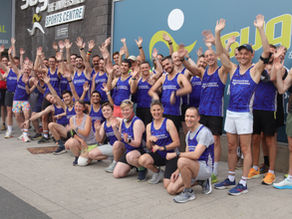 Members of a running club, most wearing blue vests with the name "MANCHESTER FRONTRUNNERS" printed on it in white, pose with jazz hands in front of a leisure centre, light summer evening.