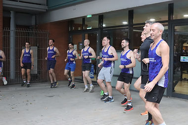 A group of runners, most wearing blue and white vests with words MANCHESTER FRONTRUNNERS on it, jog on the spot as part of a circle at a group run. Behind them is a leisure centre, with brick walls and large glass windows.