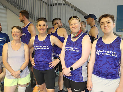 A group of women wearing running vests (three in Manchester Frontrunners blue and white vests) pose for a photo.