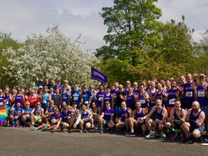 A group of runners, many wearing blue/white Manchester Frontrunners vests, pose for a group photo in a park, some holding pride flags