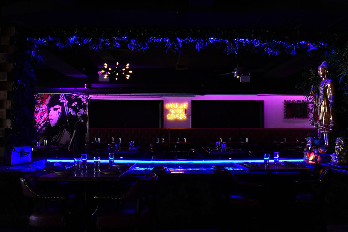 Modern dining area at Jade Eatery & Lounge in Forest Hills, Queens with neon “Indulge Your Senses” sign and Buddha décor