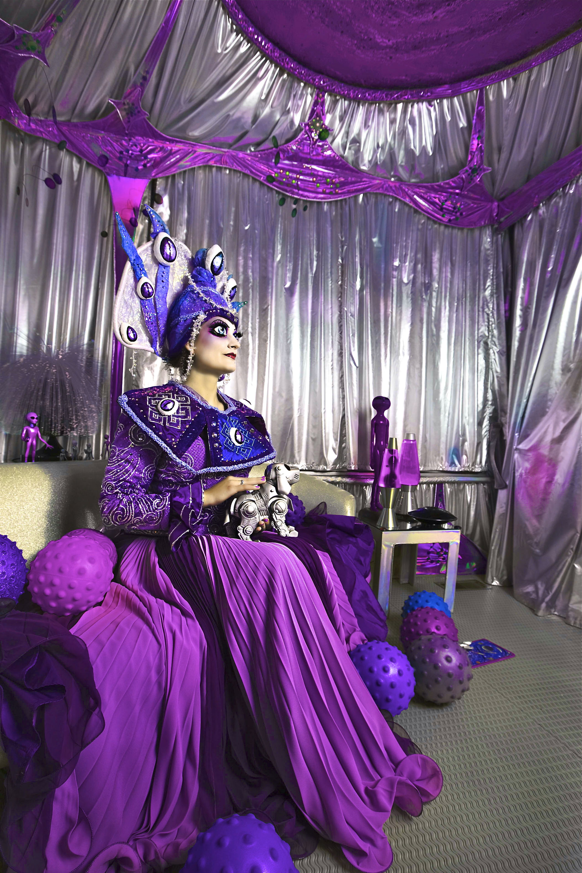 A person in an ornate purple costume with a headdress sits on a couch in a metallic room, holding a robot. Colorful balls on the floor.