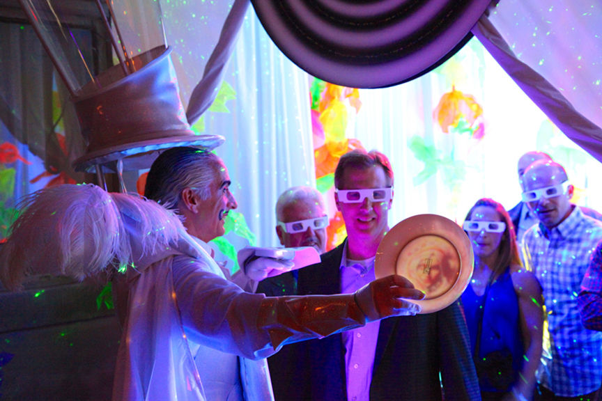 A man in a white coat and hat shows a glowing plate to people wearing 3D glasses, in a colorful, luminous room with vibrant decor.