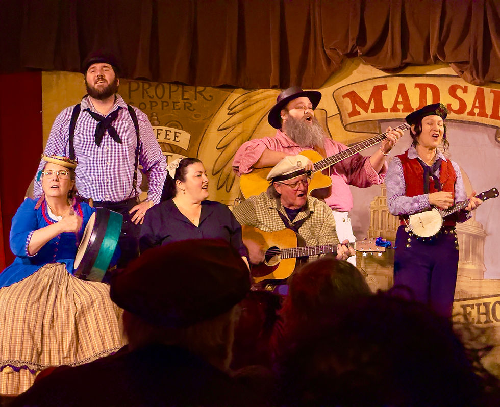 Group of six musicians in colorful outfits perform with guitars and banjos on stage. A rustic backdrop.