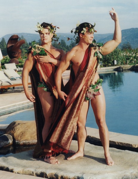 Two men in red draped outfits and leaf crowns pose poolside outdoors, with mountains in the background. They appear confident and joyful.