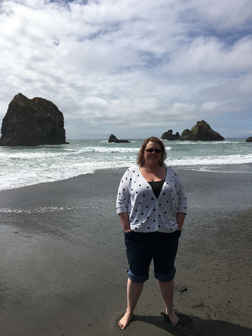 A person in a white polka-dot sweater stands barefoot on a sandy beach with rocky formations and cloudy sky in the background.