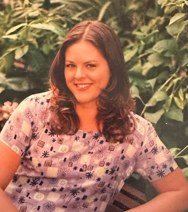 Smiling woman in a patterned purple dress sits in a lush, green garden setting.