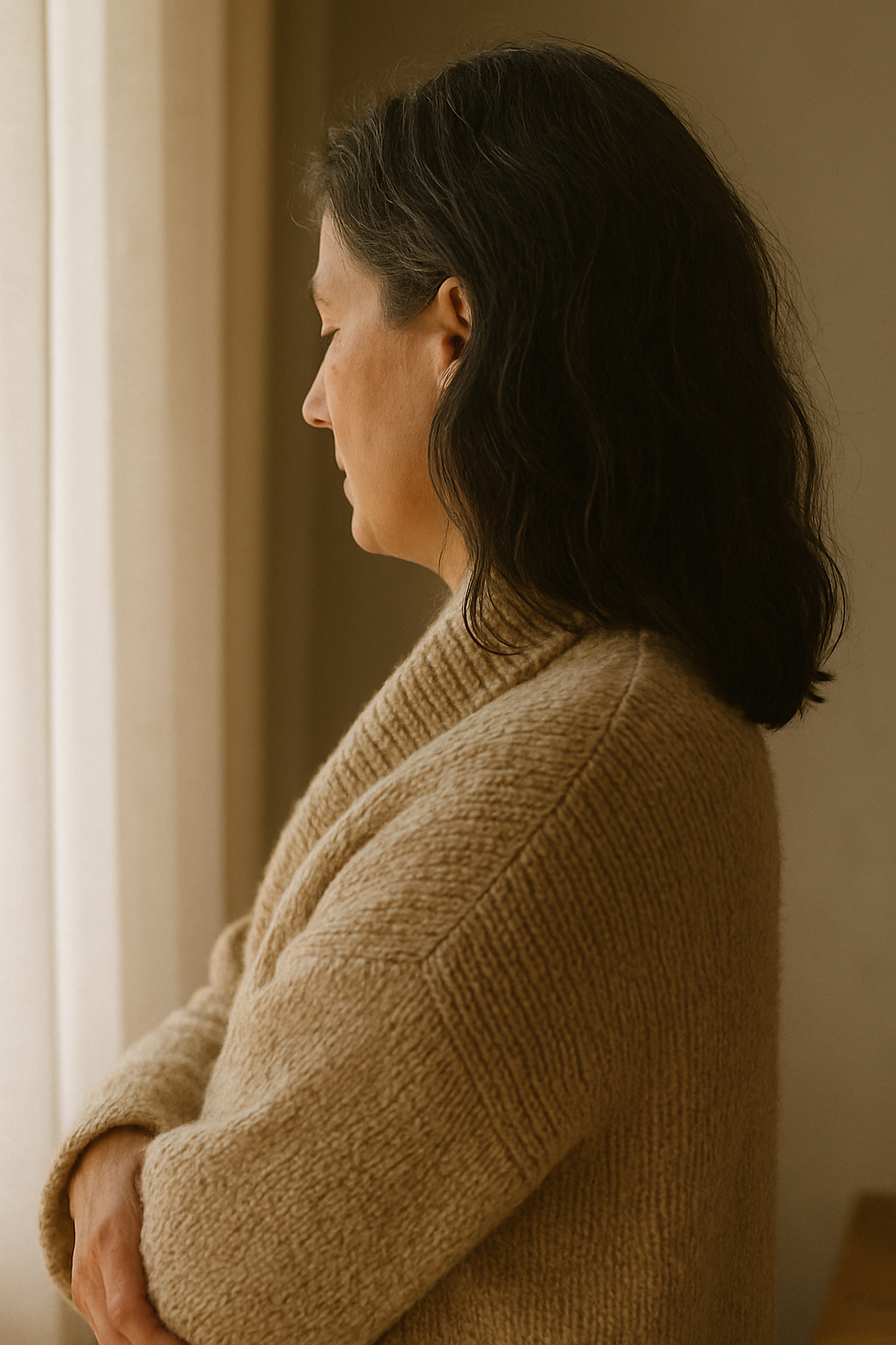 Woman in a beige sweater gazes out a window, embracing a calm and contemplative mood within a softly lit room.