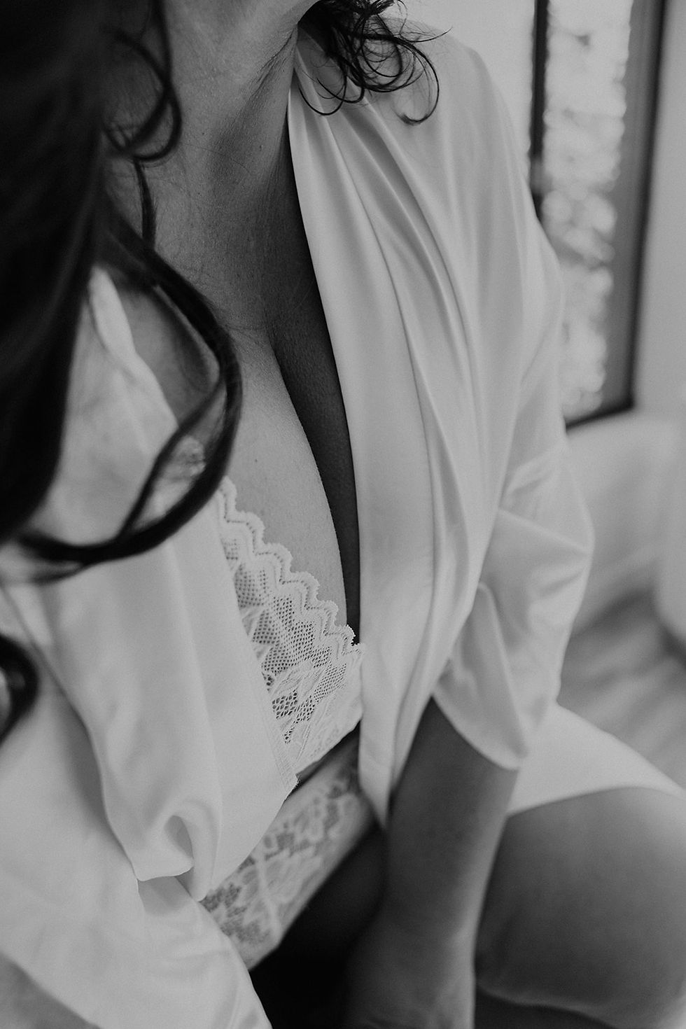 Close-up of a person in lace lingerie and a robe sitting near a window. The image is black and white, highlighting delicate fabric details.