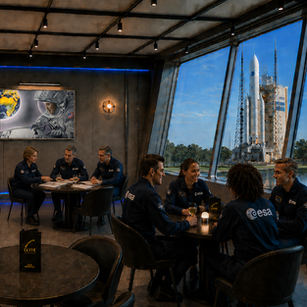 Bar moderne “Gravity” avec vue sur une fusée Ariane prête au lancement.
Des astronautes de l’ESA discutent autour d’un verre, sous le tableau “Capsule” représentant un astronaute observant la Terre depuis l’espace.