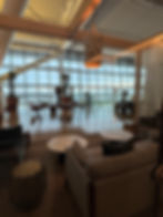 Cozy lounge with sofas, round tables, and plants, overlooking an airport runway through large glass windows. Soft lighting sets a calm mood.