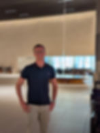 Sam smiling in a spacious, modern lobby with beige walls, a large light fixture, and a blue-lit window in the background.