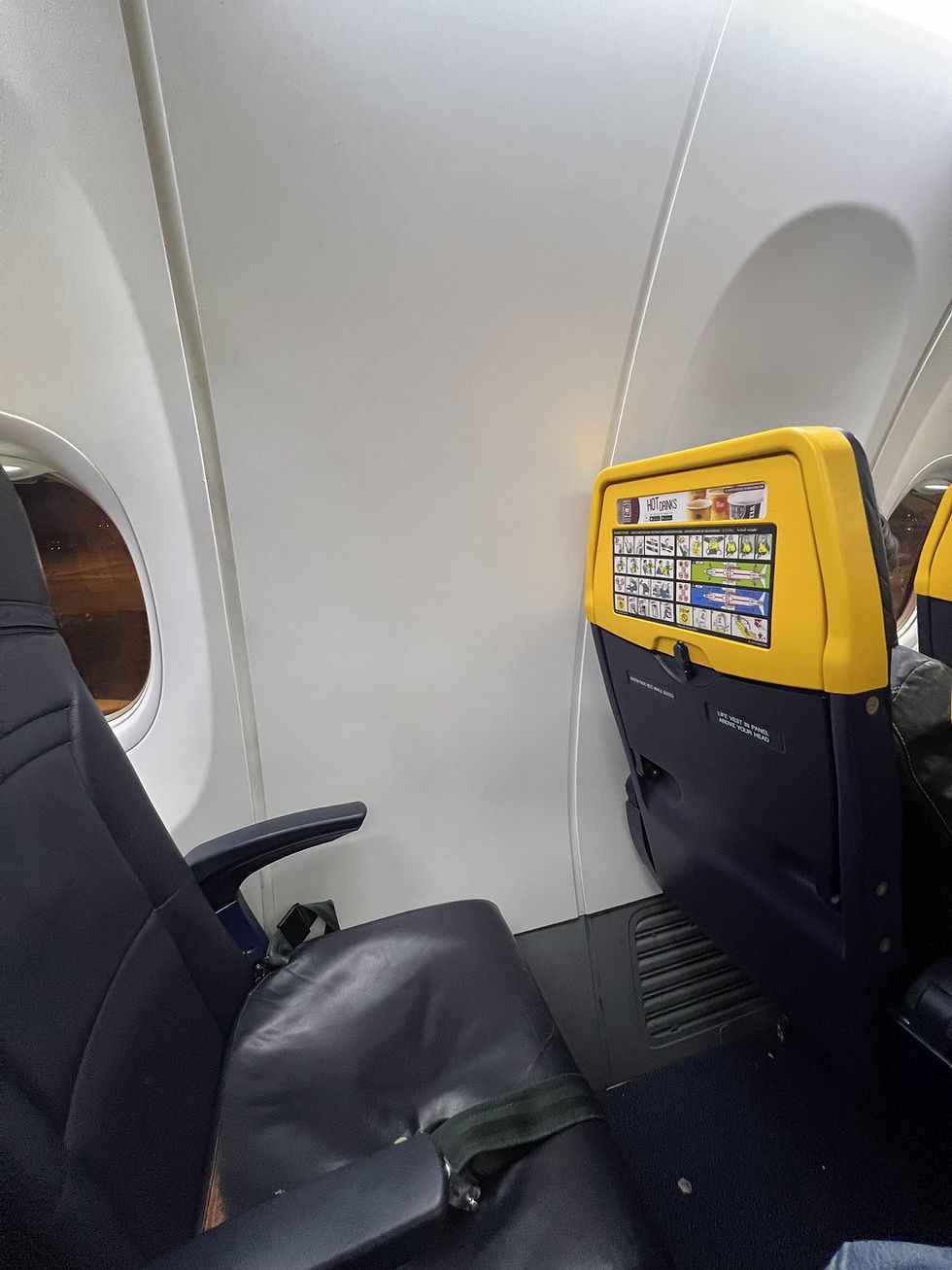 Airplane interior showing an empty dark blue seat beside a window. The seatback ahead has a yellow top with safety instructions.
