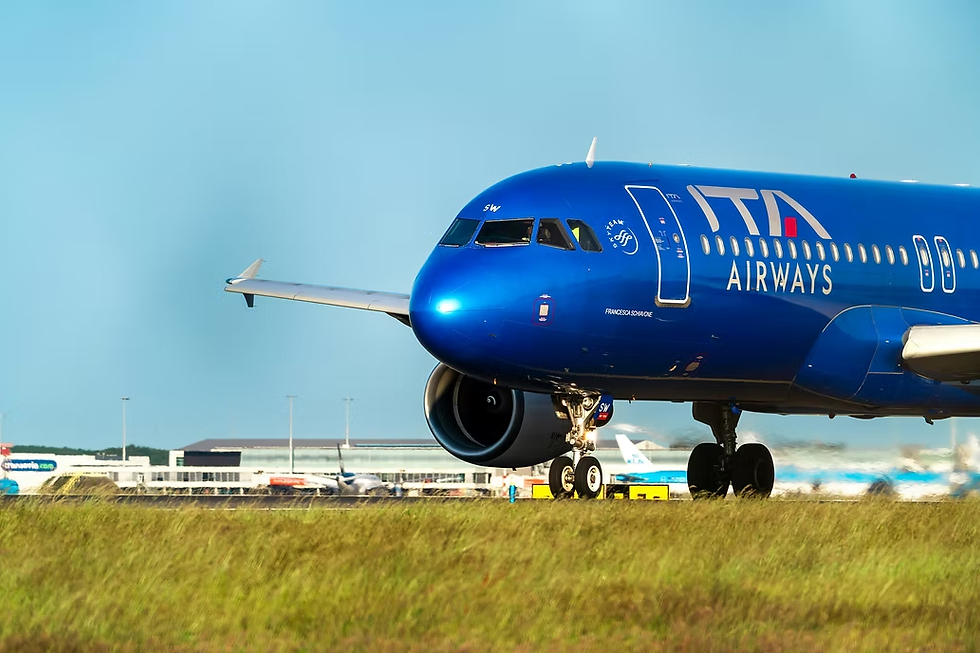 Blue ITA Airways plane taxies on grassy runway, with airport in the background under a clear sky. Text: ITA Airways.