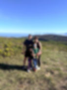 Smiling family of four stands on a grassy coastal hill with ocean and mountains under a clear blue sky.