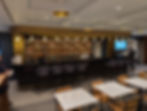Modern bar with gold accents and overhead lights. Black chairs at counter, wooden tables in foreground. A person stands by the bar.