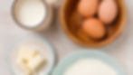 Eggs in a wooden bowl, flour, milk, sugar, butter, oil, and shredded coconut arranged on a light wooden table for baking preparation.