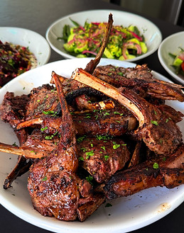 A plate of grilled lamb chops sprinkled with fresh parsley, with three bowls of salad in the background