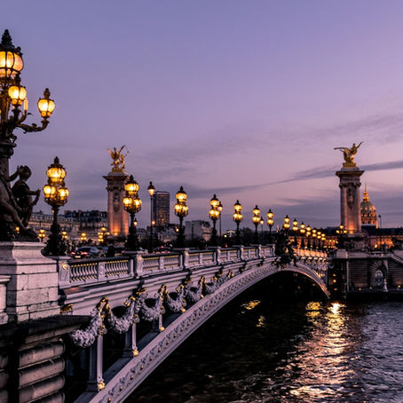 The Pont Alexandre III bridge in Paris at twilight, featuring ornate golden statues and street lamps reflecting over the Seine River with the Eiffel Tower silhouette in the distance.