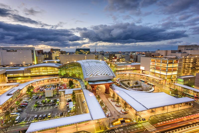 Skyline of a city at dusk, featuring a brightly lit, modern train station with snow-covered roofs. A vibrant, lively urban scene.