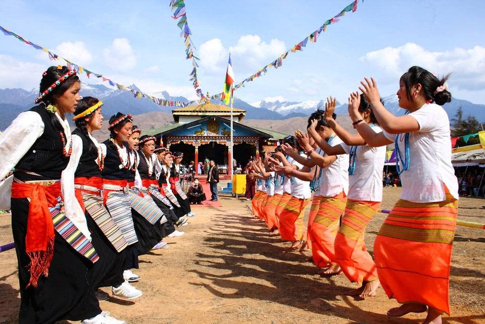 Women in traditional dress dance facing each other at a mountain temple, with prayer flags and snowy peaks under a blue sky.