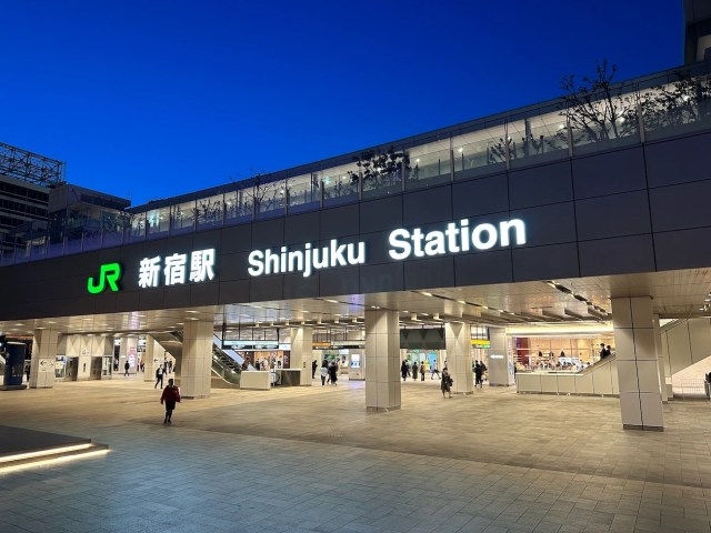 Shinjuku Station entrance at dusk with illuminated signs. People walk in a modern plaza under a deep blue sky. Mood is calm and urban.