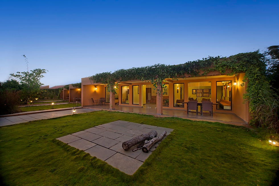 Dusk view of a modern villa with warm-lit patio, vine-covered roof, manicured lawn, and a stone fire pit under a clear blue sky