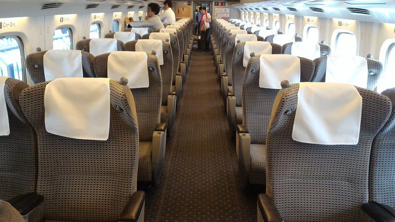 Empty train with patterned seats and white headrests, long aisle, two people walking at the front. Bright windows lining the walls.
