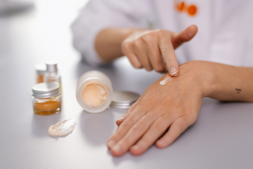 Hands applying cream from a jar on a table, with small containers nearby. Soft lighting, neutral colors, calm setting.