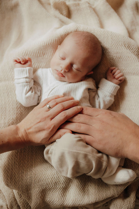 Neugeborenes Baby liegt schlafend auf beiger Decke.
Sieht sehr entspannt aus, da die Arme neben dem Köpfchen liegen. Mama und Papa haben ihre Hände sanft auf dem Bauch des Babys liegen. Sehr sanftes und liebevoll, harmonisches Bild.