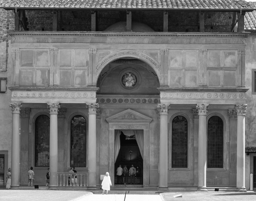 Classical-style building façade with central arched entrance, flanked by columns and topped with a circular relief; upper panels feature decorative motifs, with people standing near the doorway.