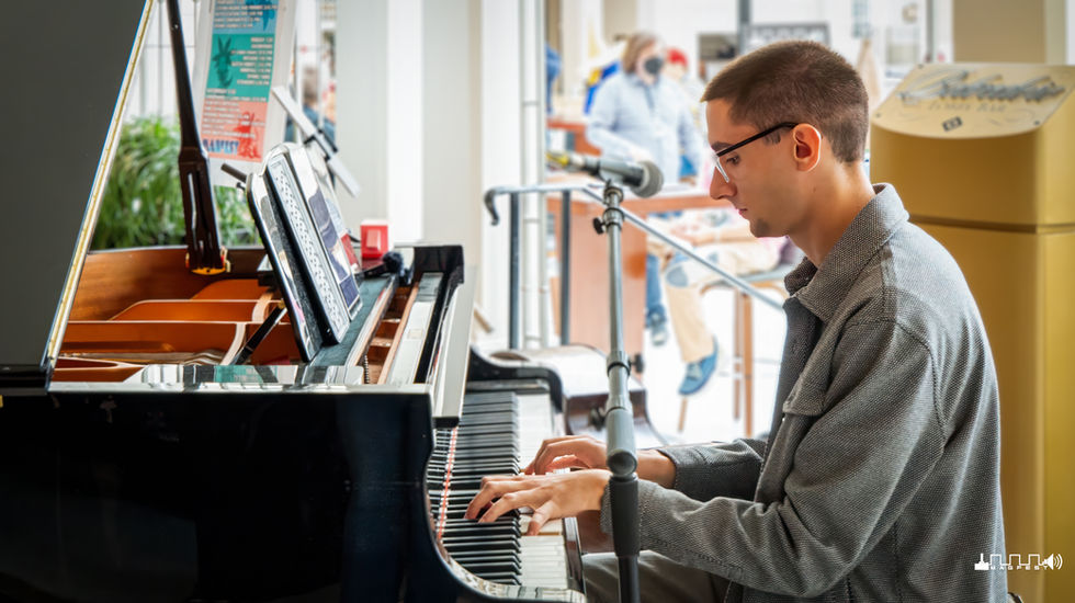 Person playing a grand piano indoors at MAGFest with the lid open, revealing strings and hammers; a microphone stand and cardboard box are nearby, and other attendees—some wearing masks—are visible in the background, suggesting a public event during health precautions.